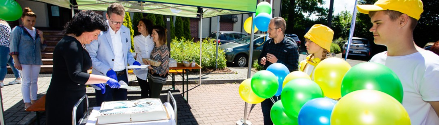 Dwie osoby kroją tort, z boku dwie osoby z balonami